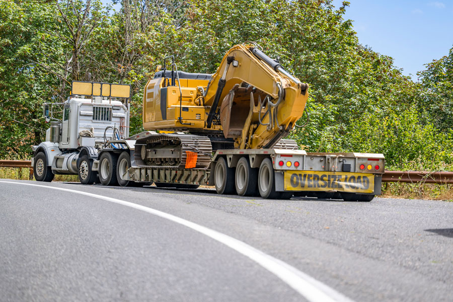 A Big Rig Semi Truck with Oversize Signs hauls a large yellow excavator on a flatbed trailer marked "OVERSIZE LOAD" along a curved, tree-lined road.